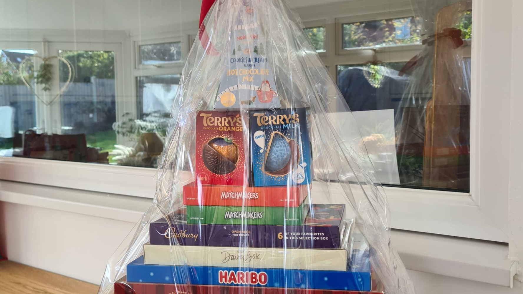 A selection of Christmas treats, from share boxes of sweets to trays of chocolates, stacked in a Christmas tree shape and wrapped in cellophane. The hamper sits in the CAP office at work as part of our Christmas fundraising.