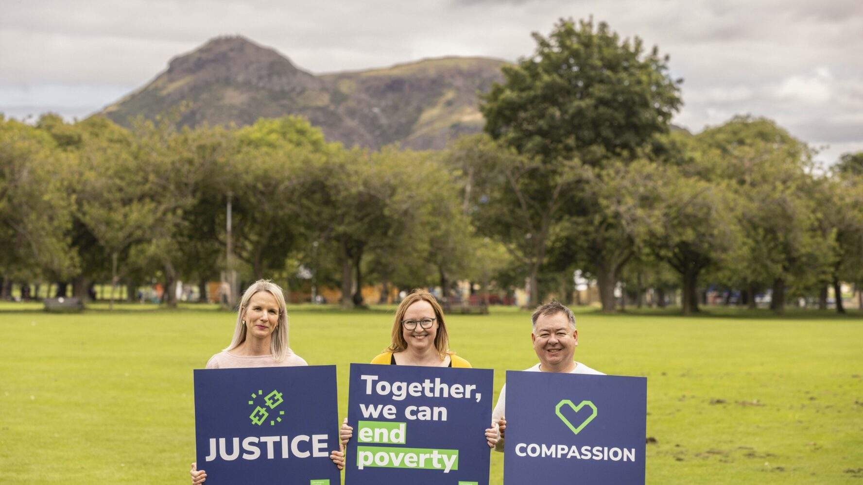 Three people stand in a field holding navy blue boards featuring the slogans 'justice', 'together we can fight poverty' and 'compassion'