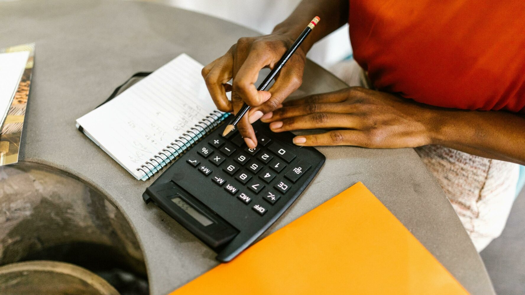 Woman typing on a calculator, holding a pen ready to write in her notebook which is next to her.
