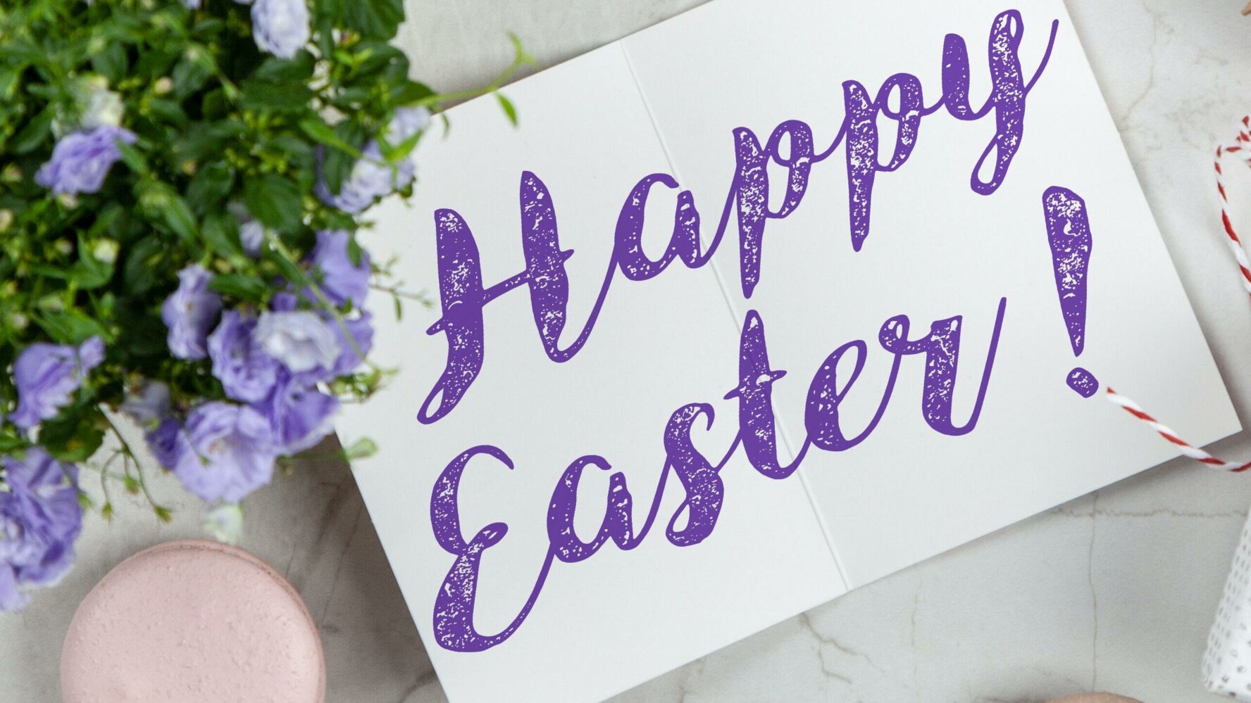 An Easter flat lay on a table with a sign with purple writing saying 'Happy Easter' and some purple flowers.