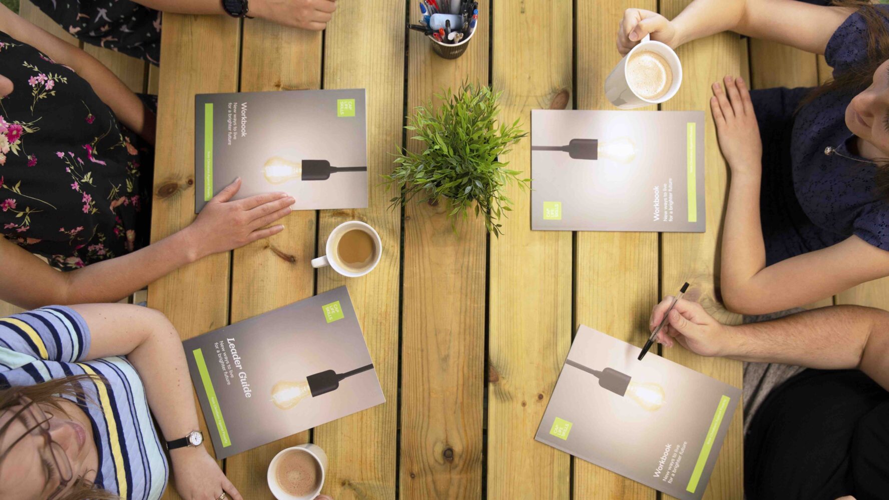 Group of men and women sat around a table holding cups of coffee and looking at workbook leaflets.