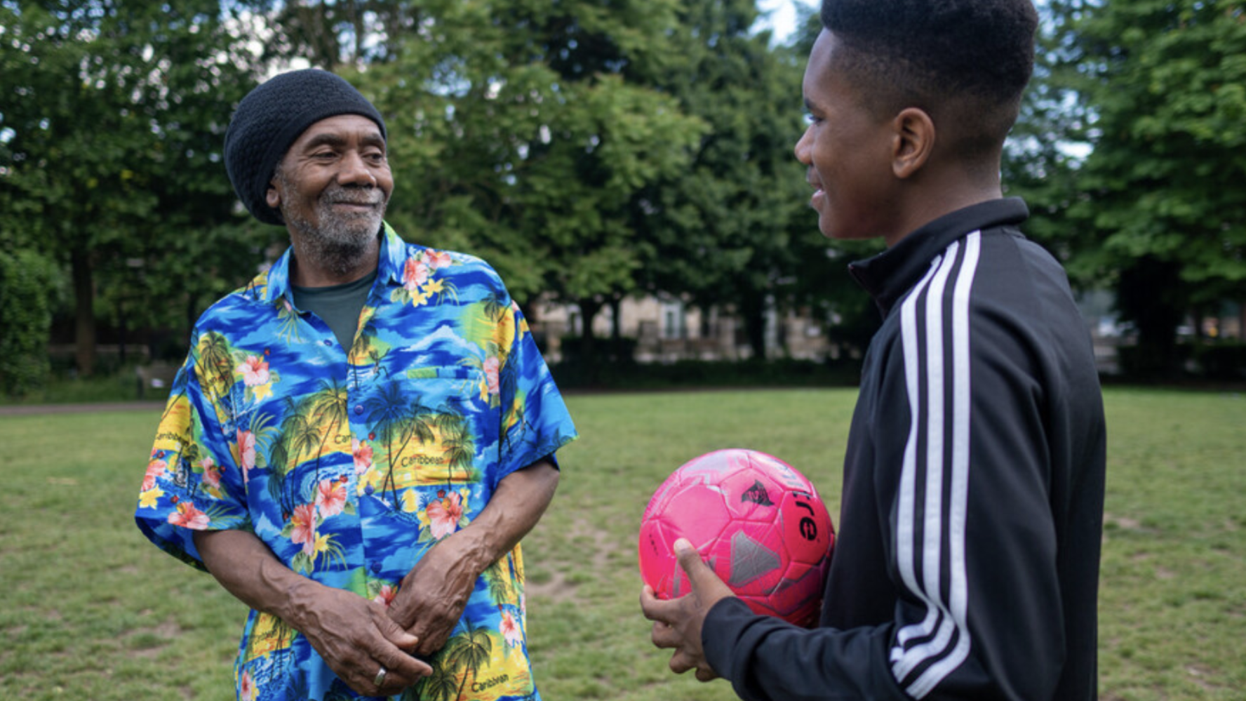 Image of a father and son smiling at each other and outside in a park, the son is holding a football