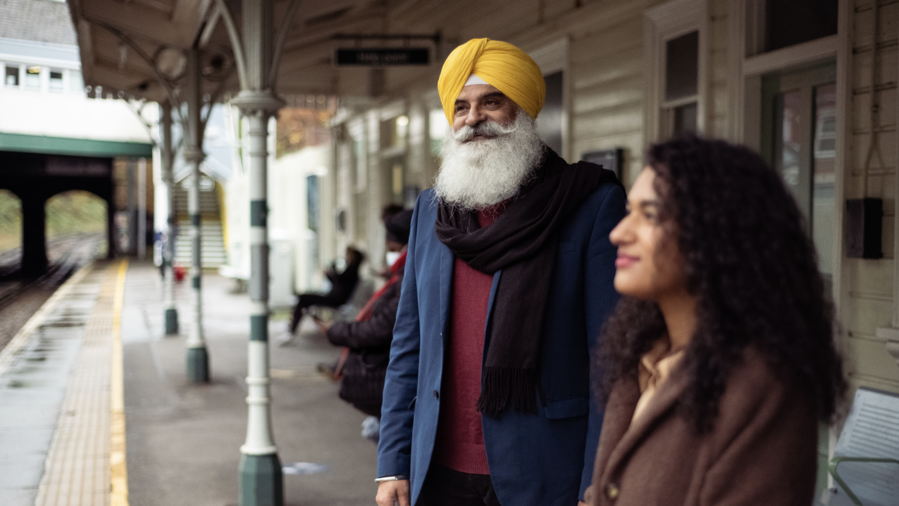 A man and a woman stood on a UK train platform, smiling and waiting for their train