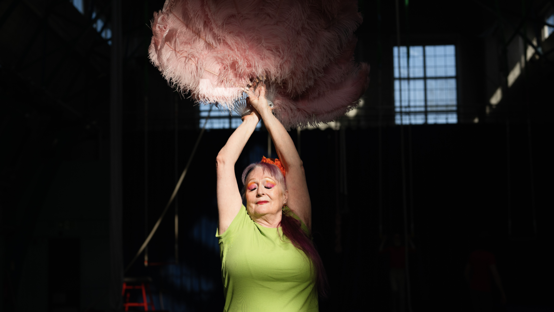 An older lady on stage holding a feather boa in the air and wearing bright clothing