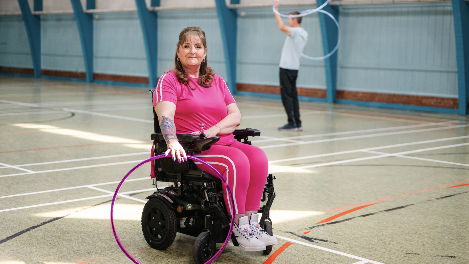 A lady in a wheelchair wearing pink athletic clothing, at her local gym holding a hulahoop.