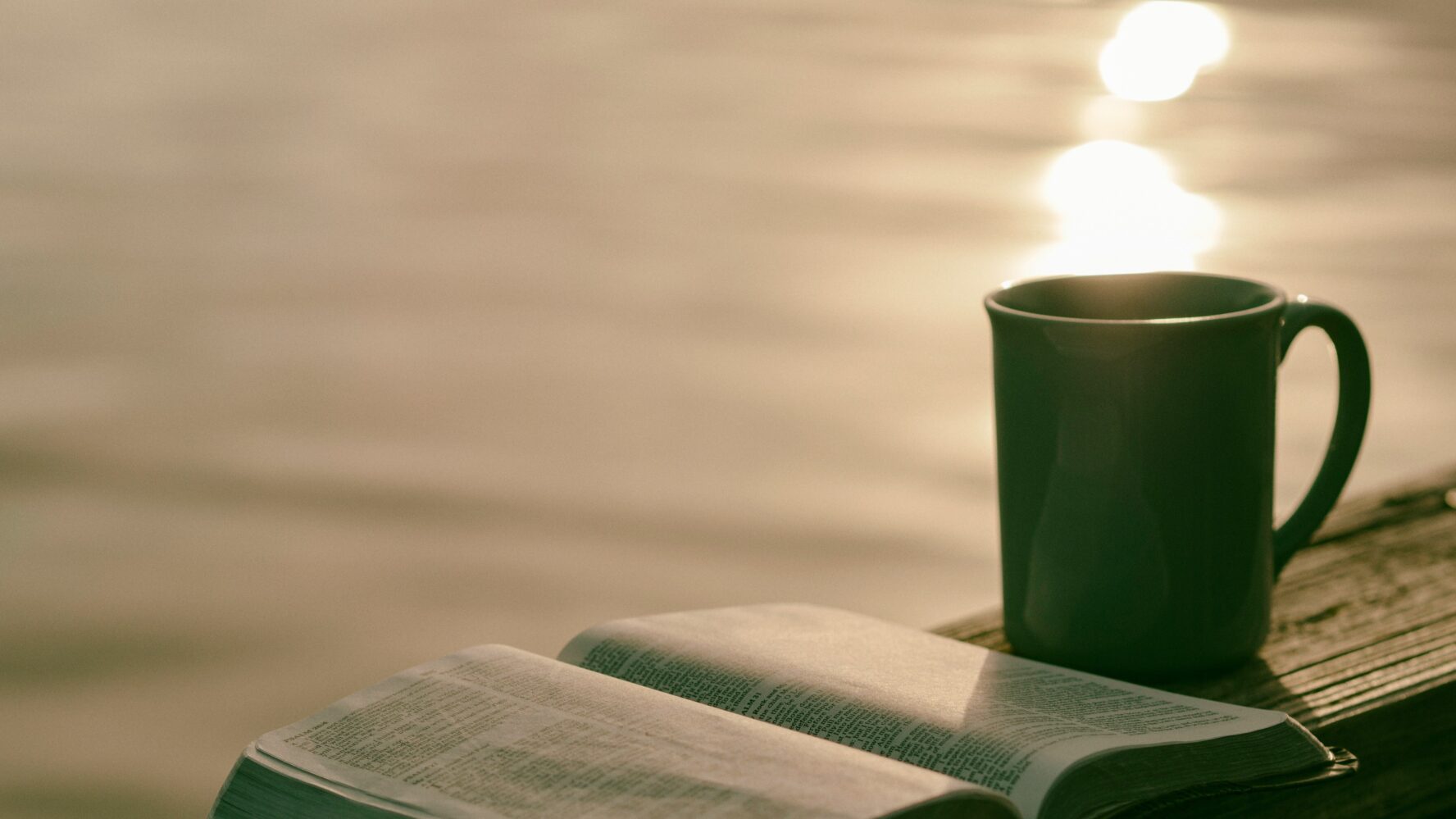 An open Bible and a mug on a bench in front of a lake, with the sun shining on the water