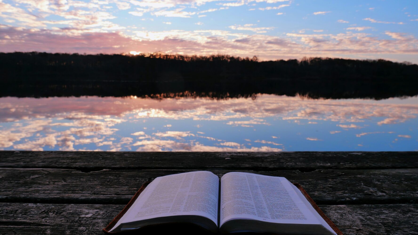 Image of an open Bible on a bench in from of a lake, with the sunset in the distance