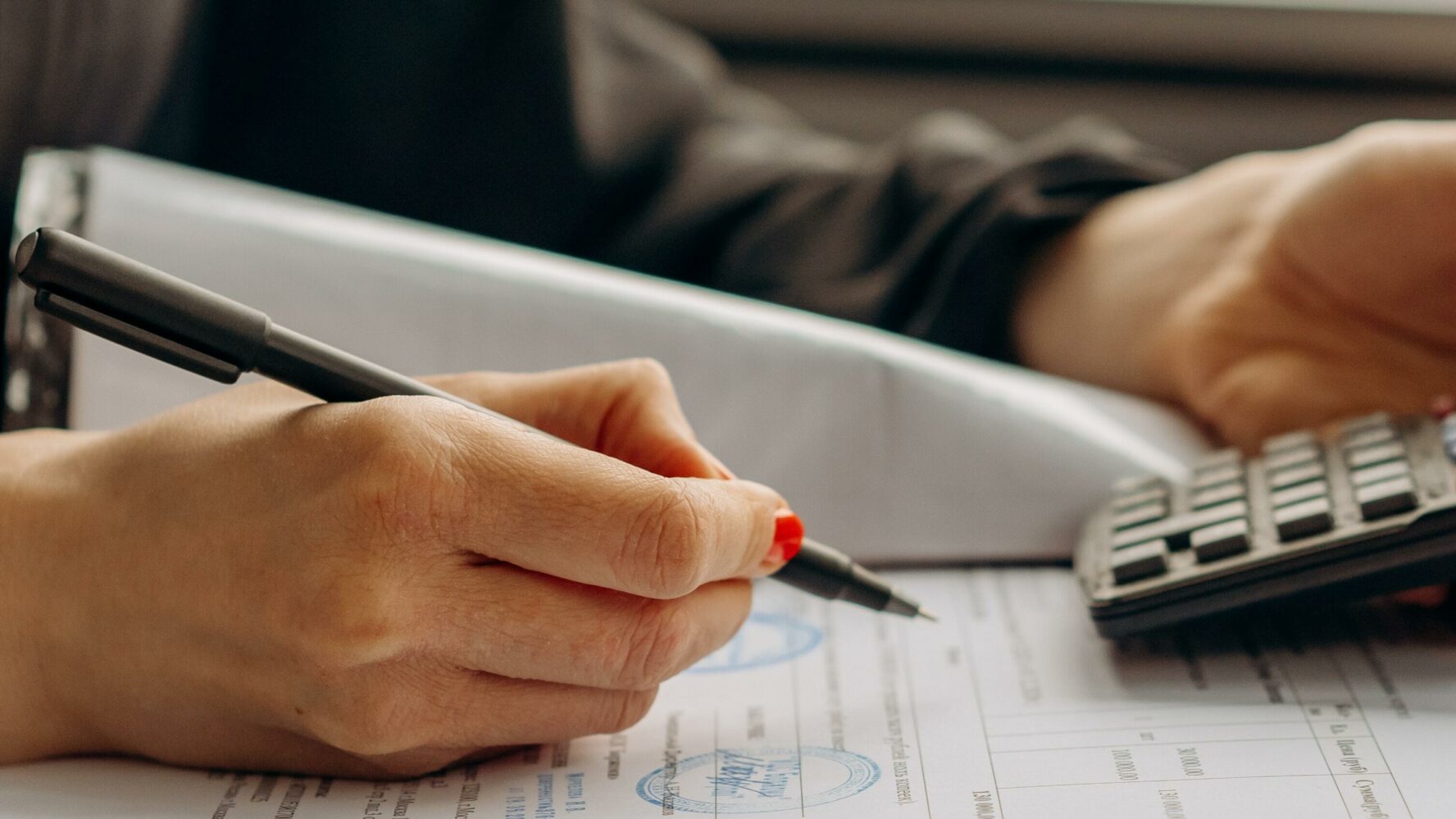 Female hand holding a pen and writing on a piece of paper, in her other hand is a calculator.