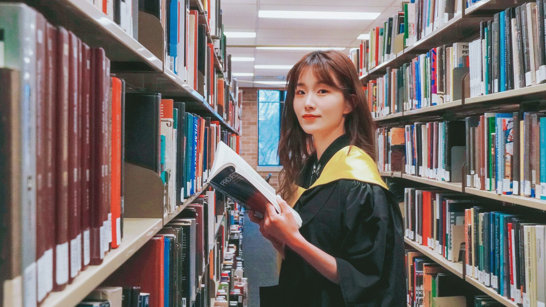 Student in graduation gown in university library, picking up a book