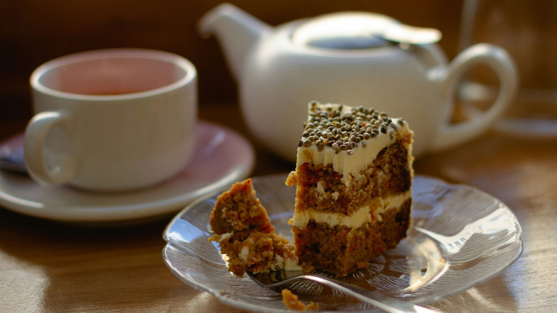 A slice of carrot cake on a plate, with a cup of tea and teapot behind it