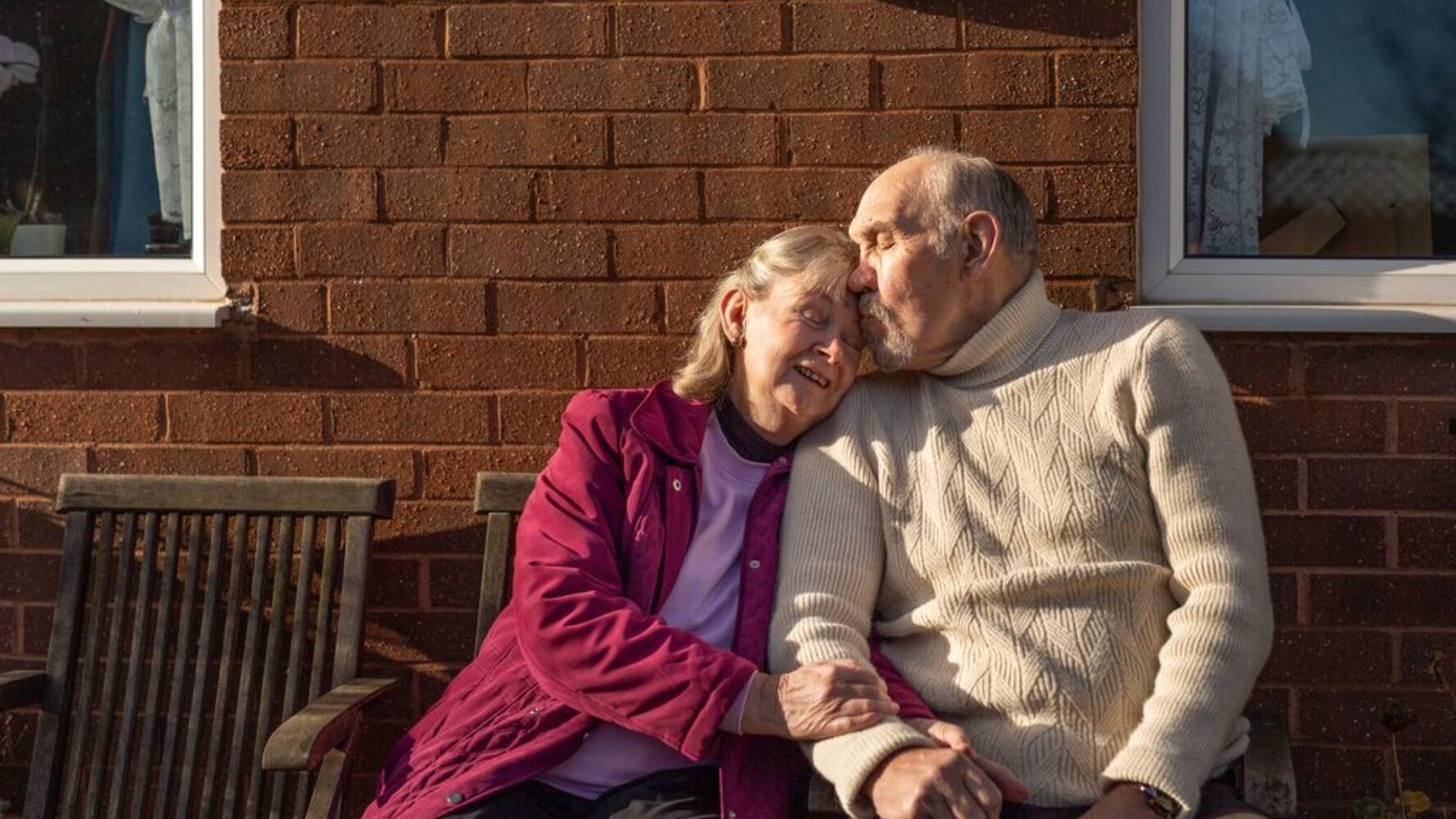 Image of older husband and wife sat next to each other, hugging on their garden bench