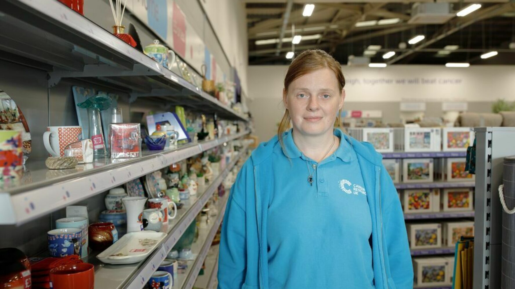 CAP client, Lyndsey, stood in a charity shop where she volunteers, smiling at the camera