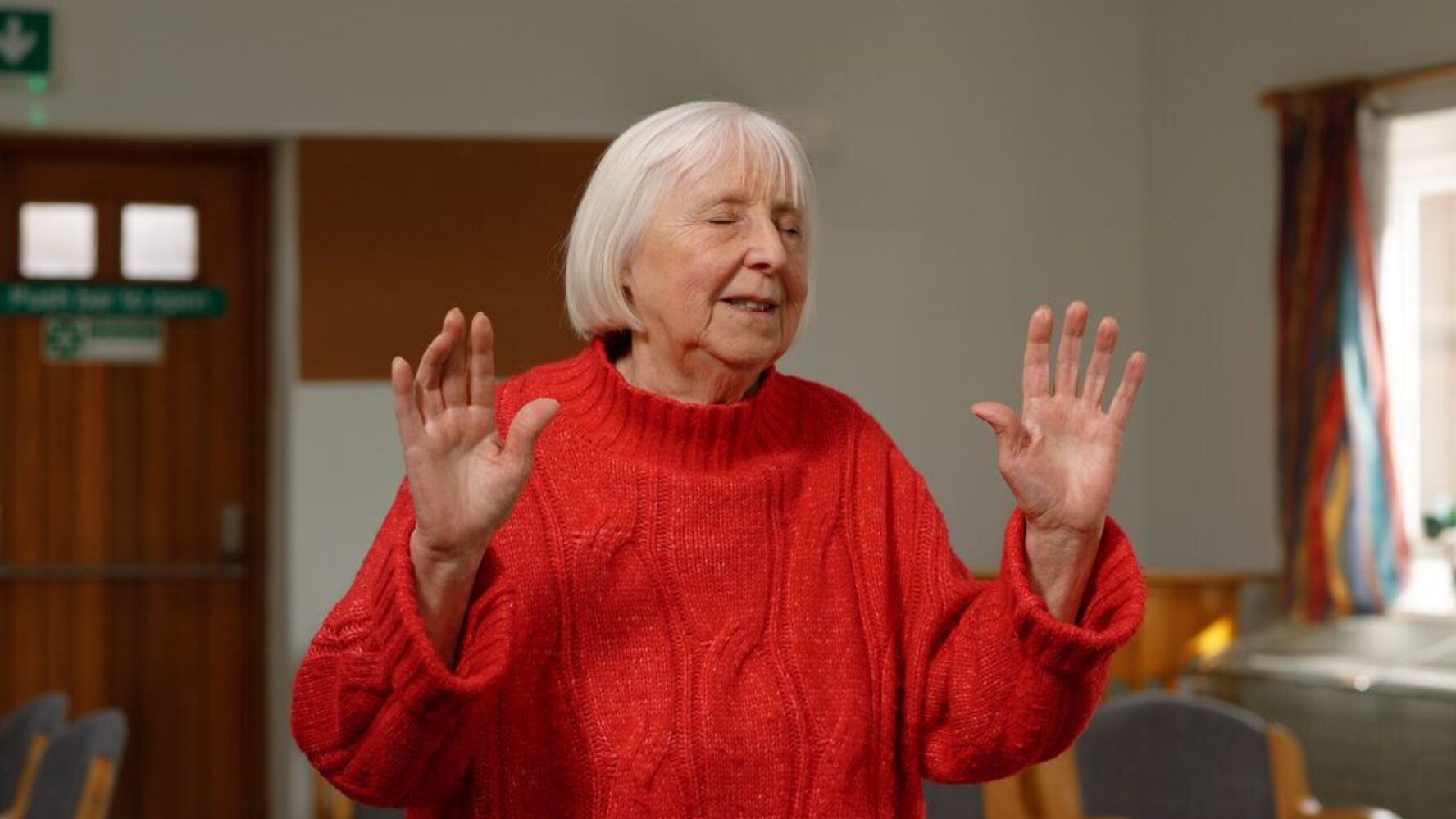 An older lady in her local church, worshipping with her eyes closed and arms lifted up