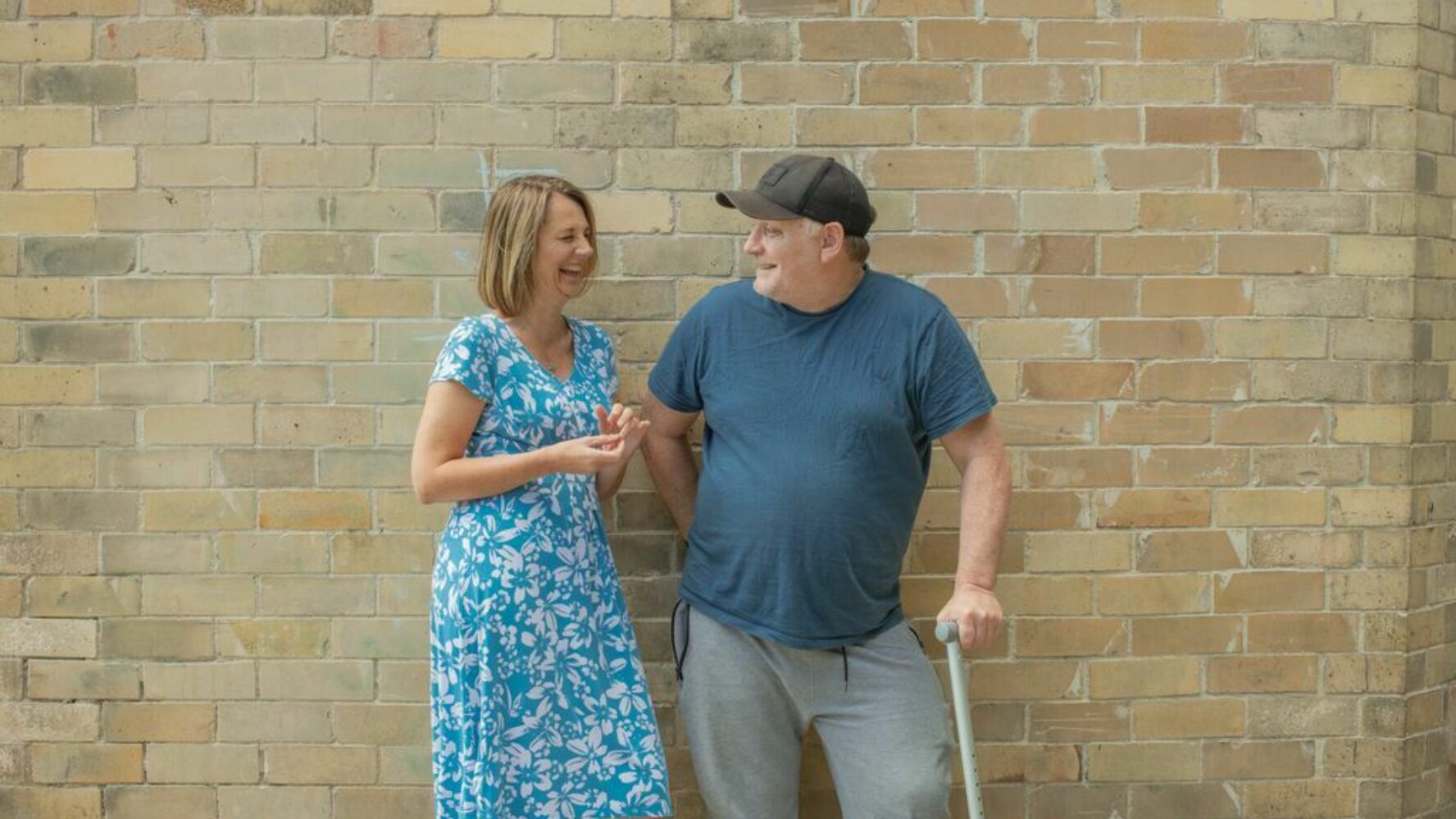 A male CAP client holding a walking stick, and his debt coach stand in front of a brick wall, both of them smiling