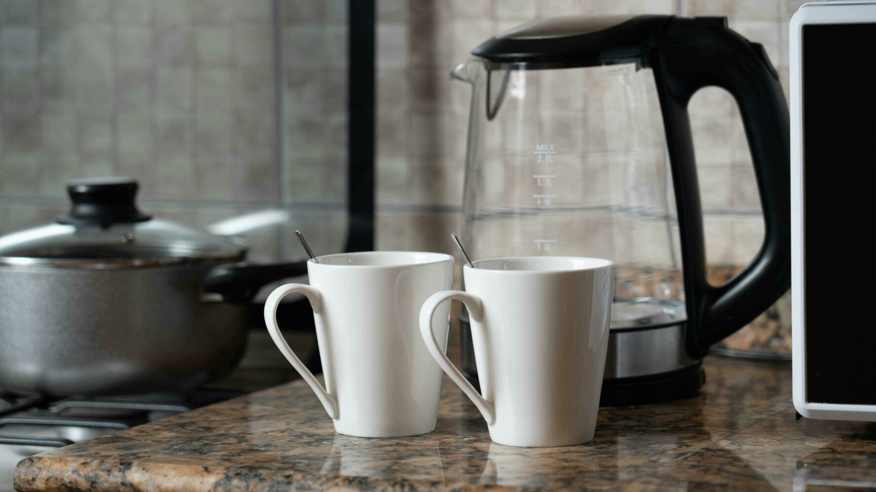 On a kitchen counter sits a clear kettle and two basic white mugs, ready for two cups of tea to be made.