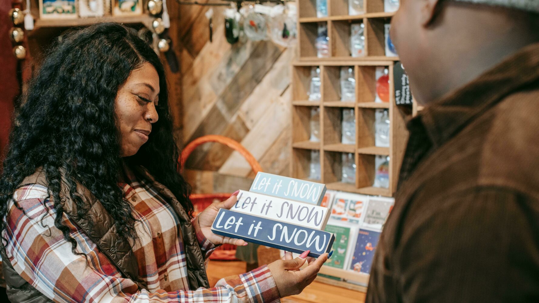 A black lady in a red and white checkered shirt is showing a black man in a brown jacket and grey wooly hat a Christmas ornament that says 'let it snow, let it snow, let it snow' as if to ask if they should buy it from the shop they're in.