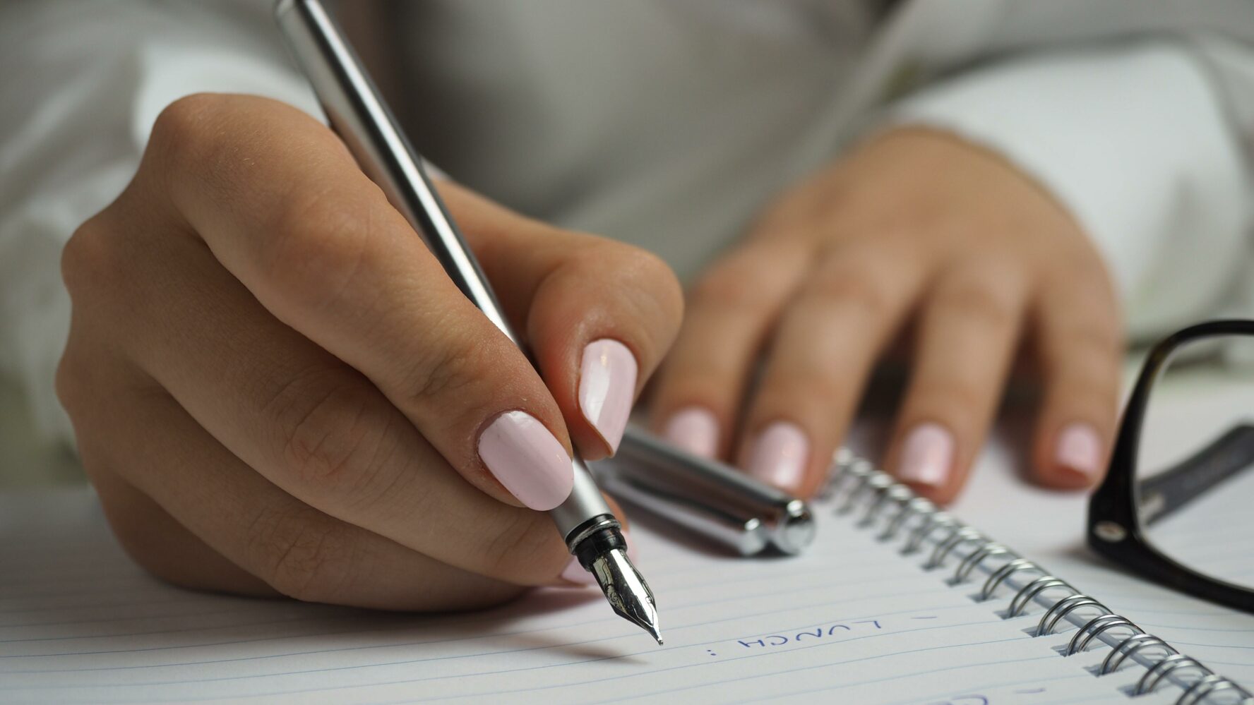 A hand with pink nail varnish holding a pen and writing in a notebook.