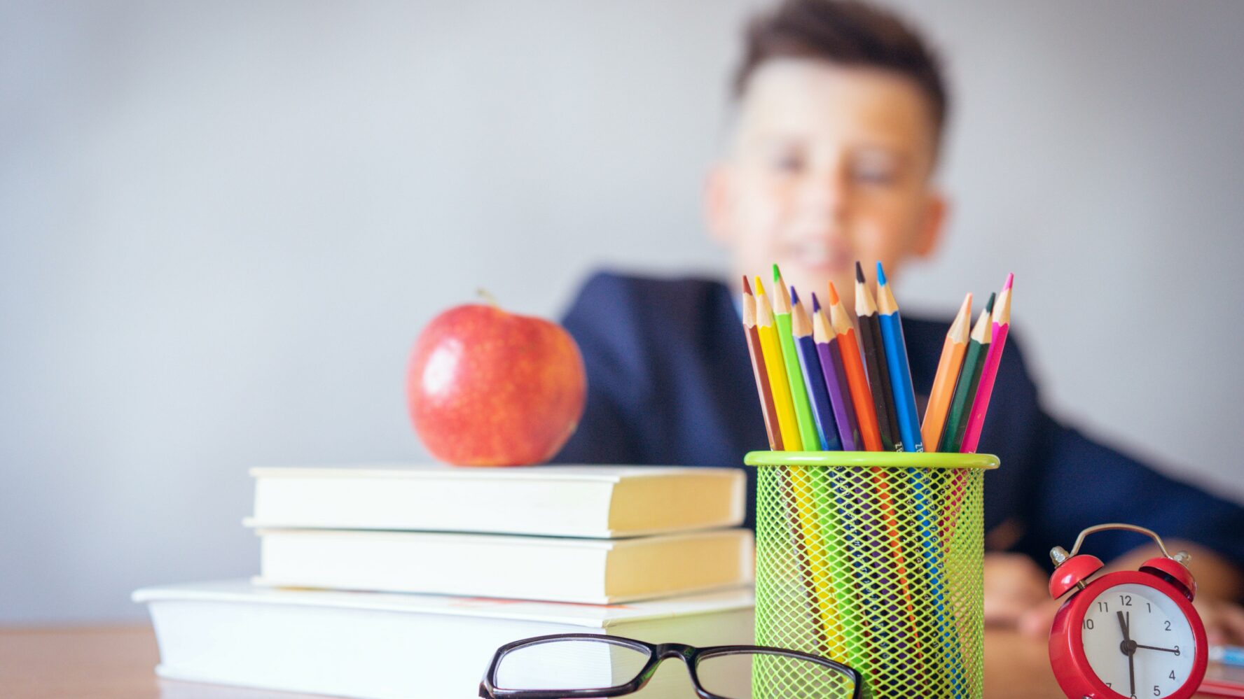 a child sits behind a pile of books, pot of pencils, apple and pair of glasses