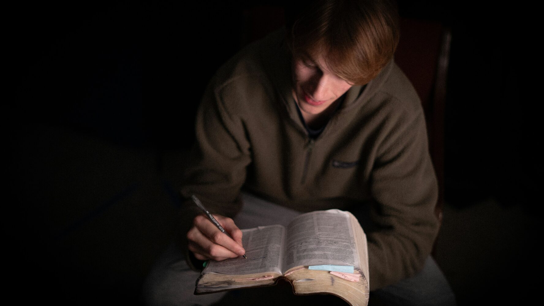 Image of a young person, sat down in the dark, looking and reading the Bible