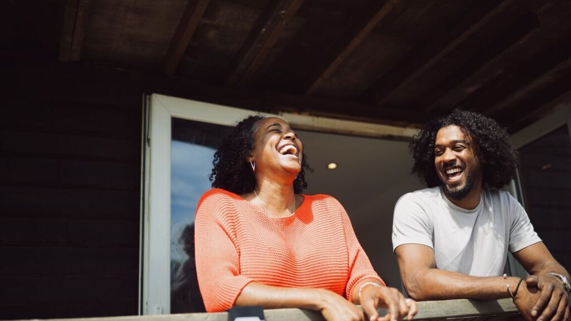 Selene, a black lady with curly dark shoulder length hair, laughing on a balcony with her brother, who is also black with curly dark hair