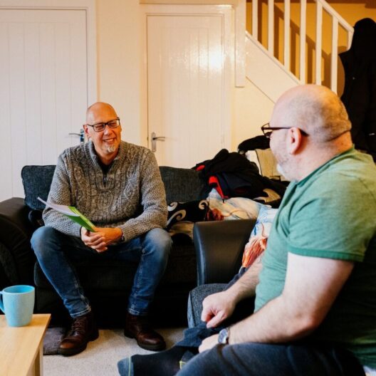 CAP client, Stevie, sat with his CAP debt coach in his living room. They are both looking at each other and smiling