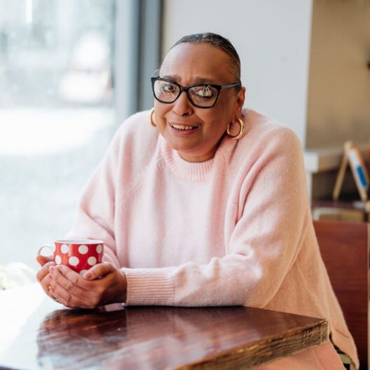 CAP client, Sharon, sat at a table holding a red mug, wearing a pink jumper and smiling at the camera