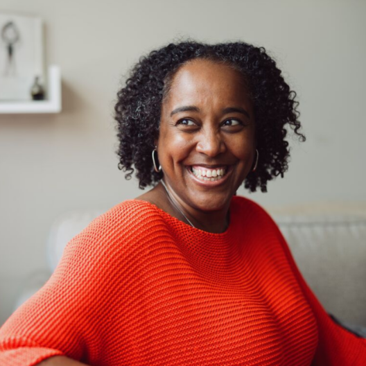 Selene, a Jamaican Indian woman with brown skin and dark curly hair. She is wearing a bright red jumper and smiling wide, as if she's laughing.