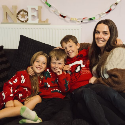Jade with her three children, Layla, Lukas and Jacob. They're sat on the sofa all cuddling together in Christmas jumpers, with decorations up behind them. They're all smiling at the camera.