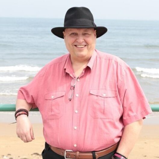 CAP client, Simon, at the seaside wearing a pink shirt and smiling at the camera, with the beach behind him.