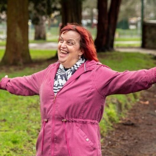 CAP client, Julie, outside in a park, smiling with her arms open wide