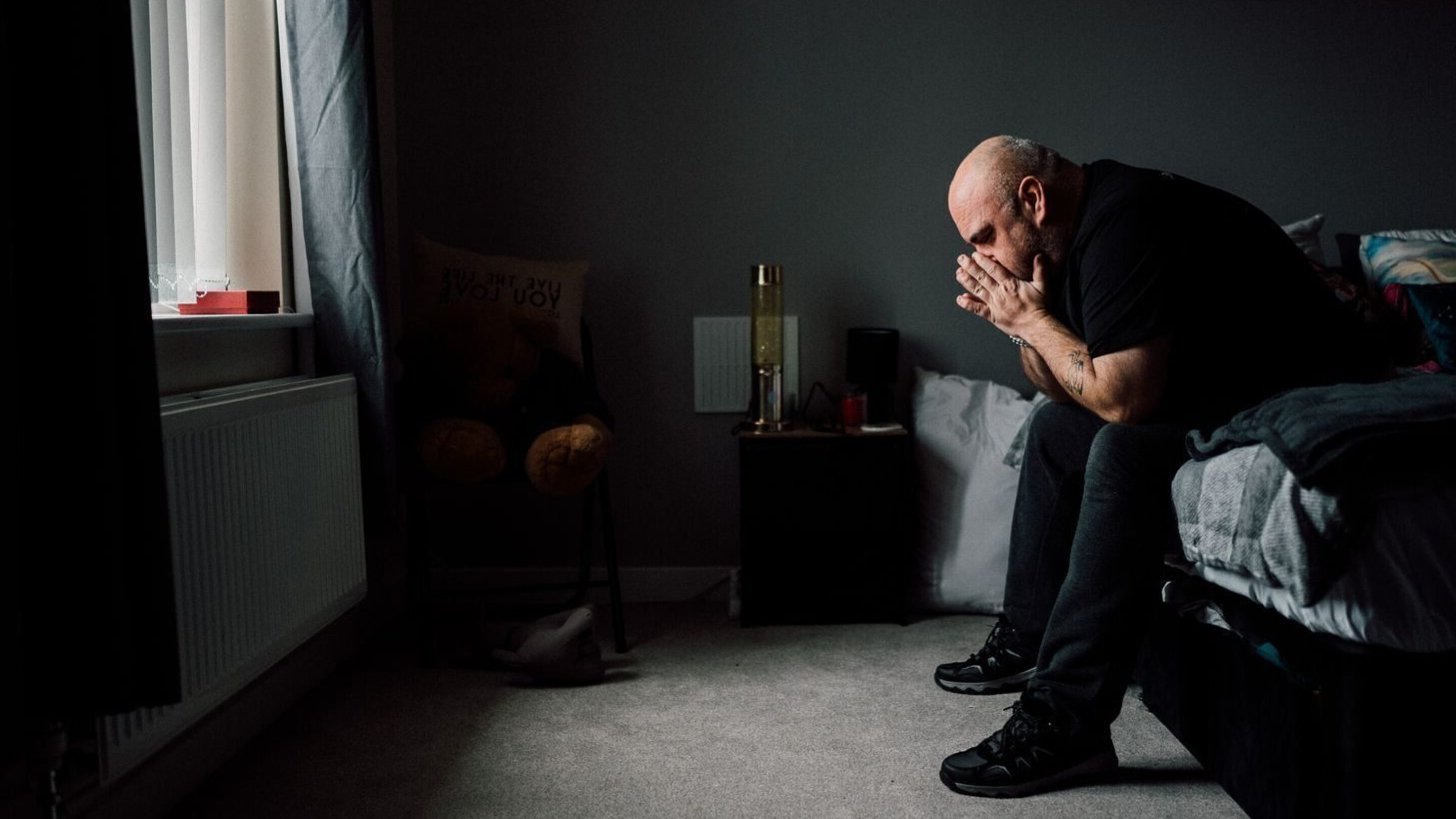 CAP client, Stevie, sat on the edge of his bed, leaning over with his face in his hands. The photo has a dark contrast to it.
