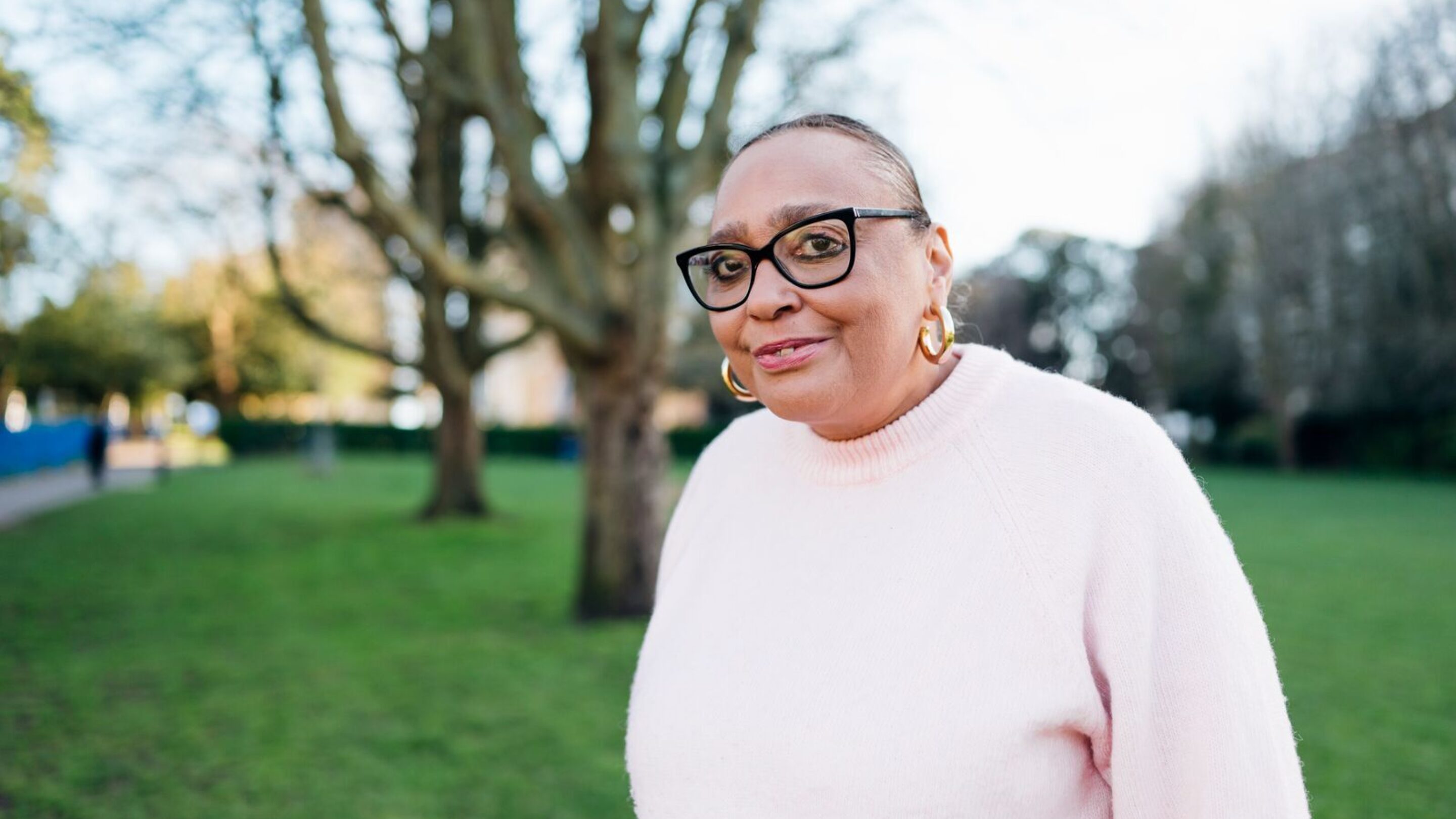 CAP client, Sharon, outside in a park, smiling at the camera and wearing a pink jumper.