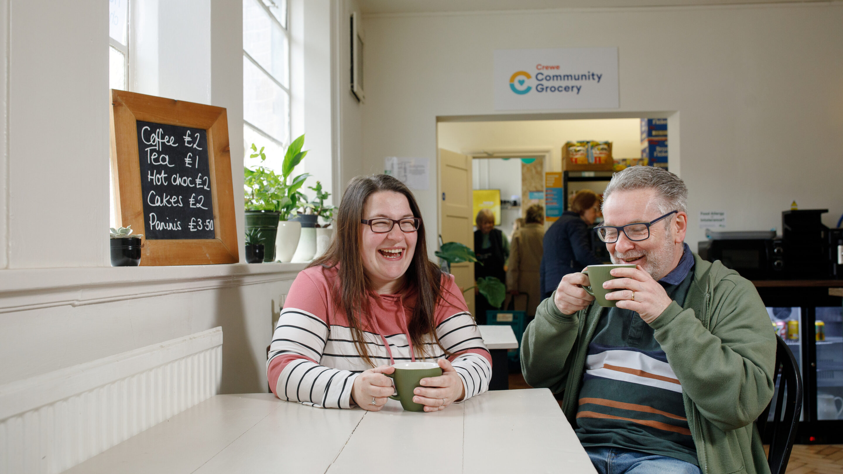 A woman with long brown hair and a man with glasses smile and drink cups of tea