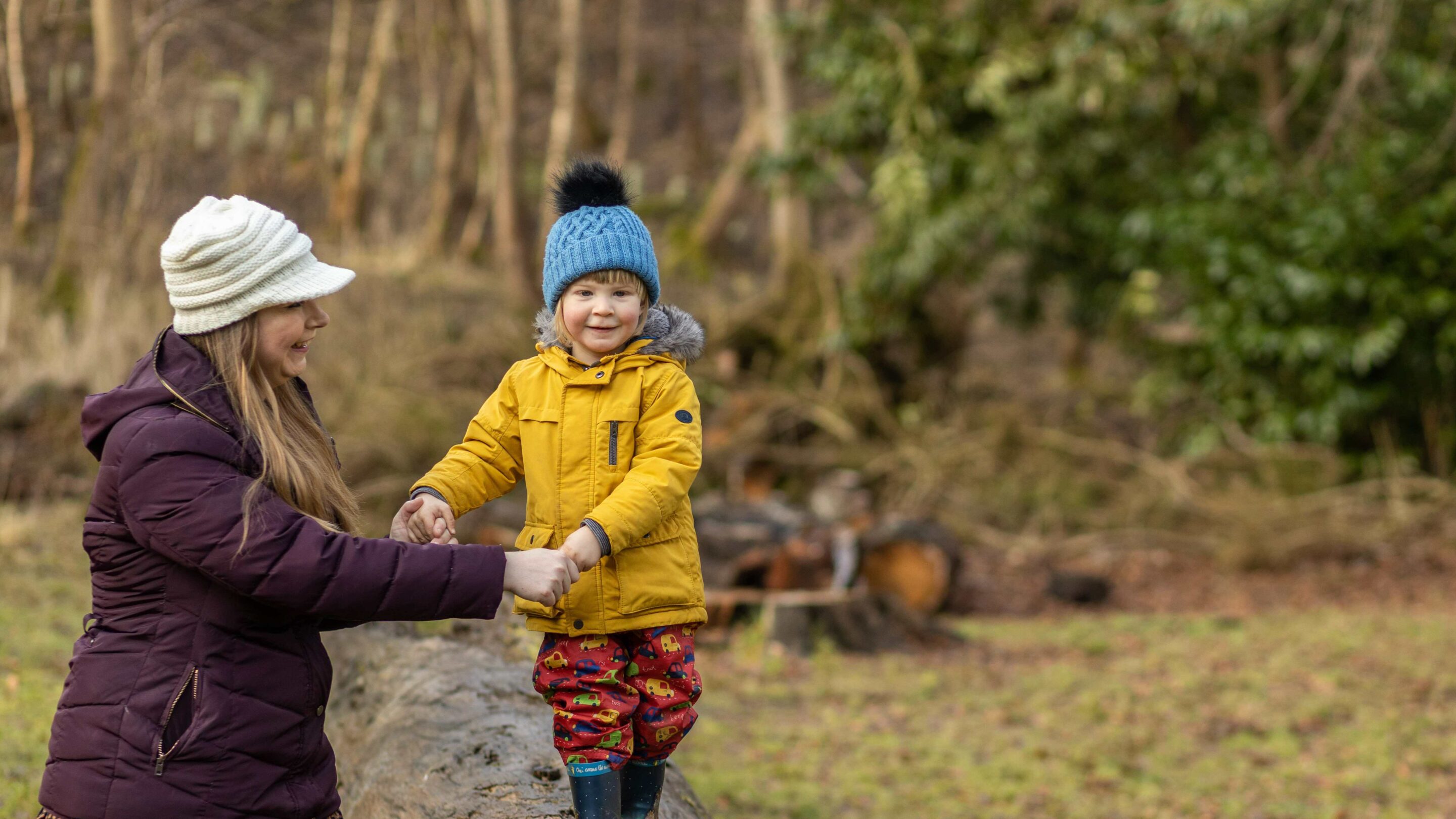 Young woman with her son playing outside and smiling, she is helping him walk along a climbing frame.