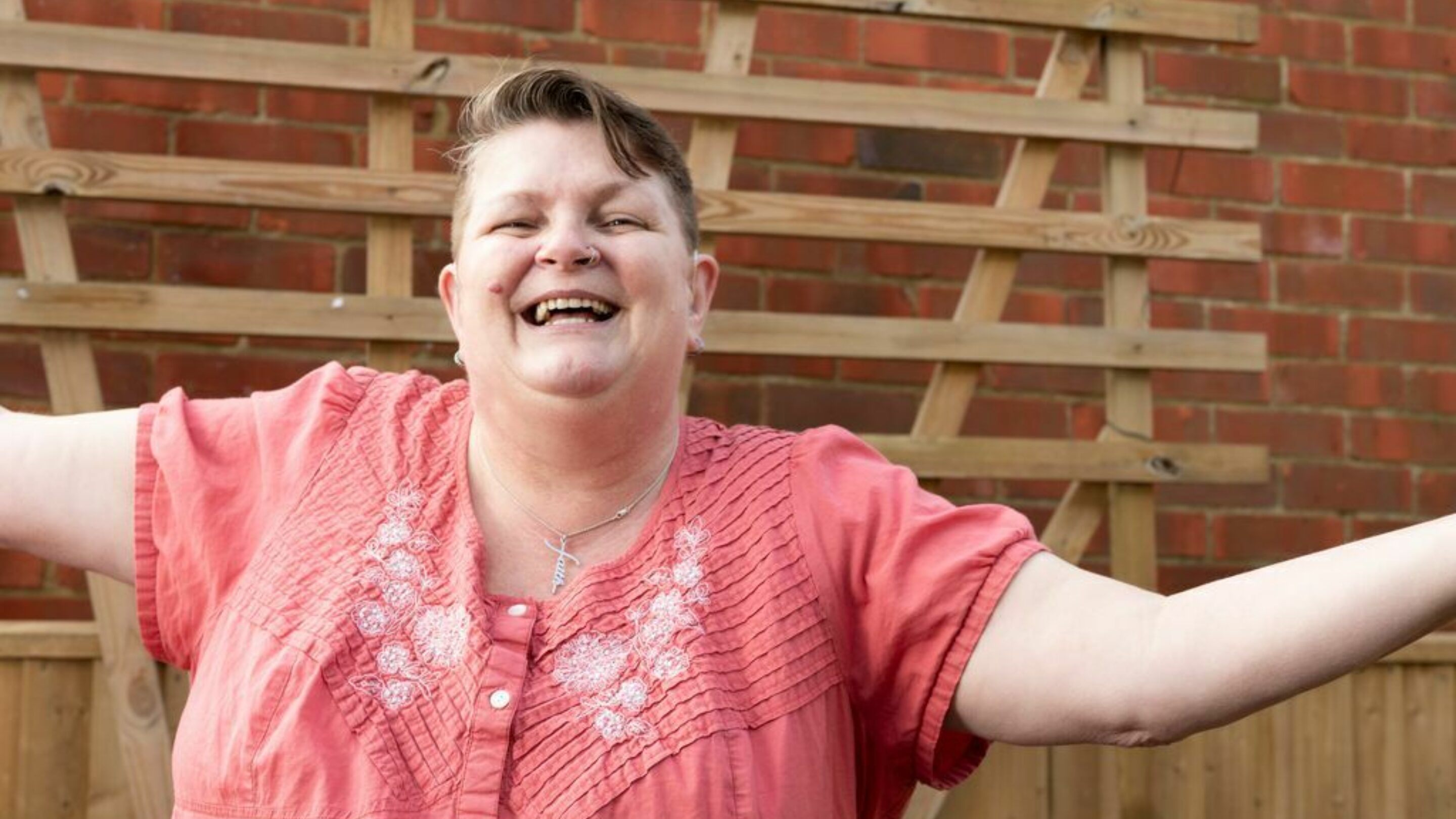 Toni, a white lady with short brown/red hair and a pink short-sleeved shirt, is a former CAP client, now debt free. She is smiling at the camera with her arms flung wide.