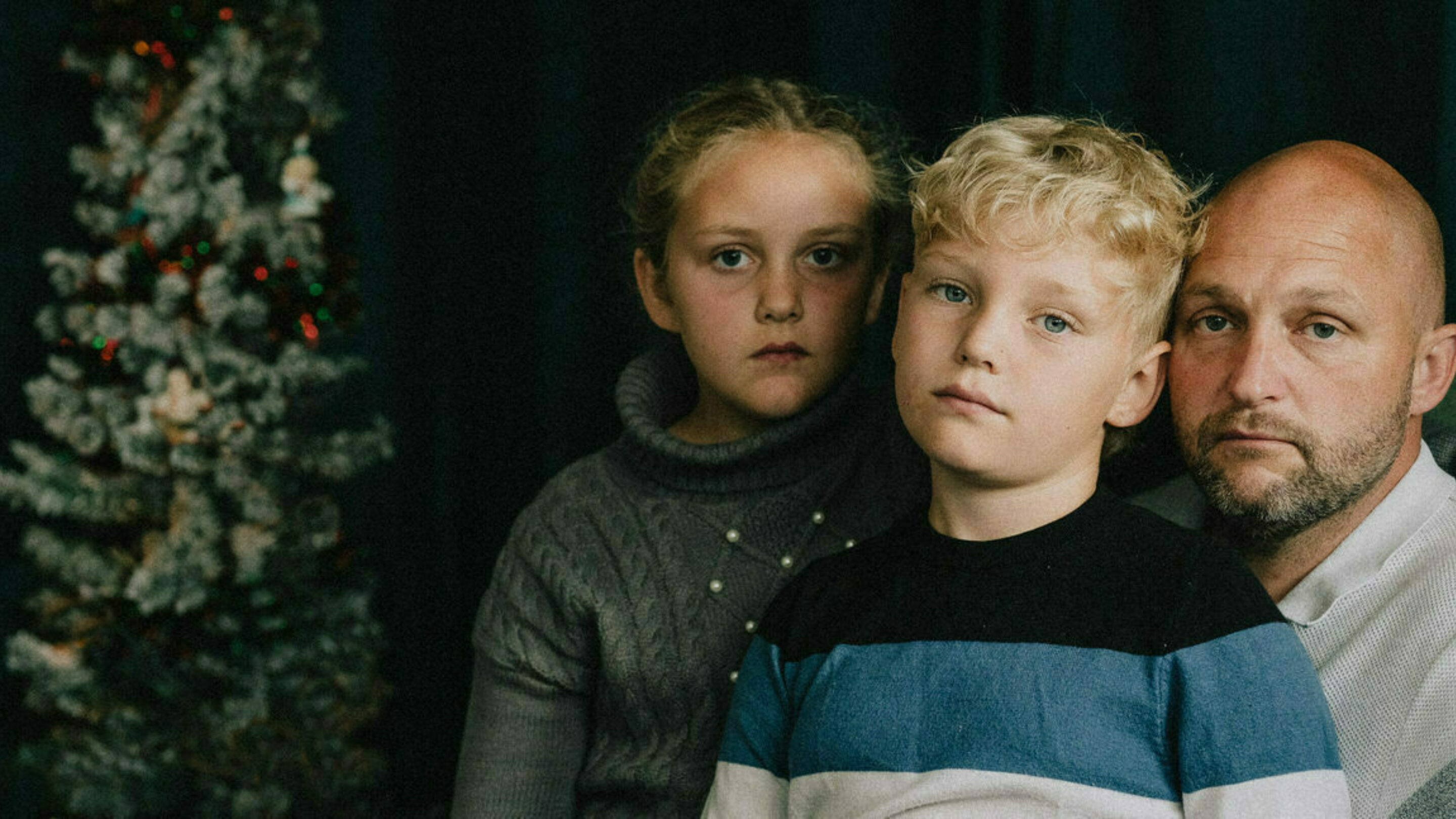 Dad sat with his two young children, all three are staring at the camera with sadness next to a Christmas tree