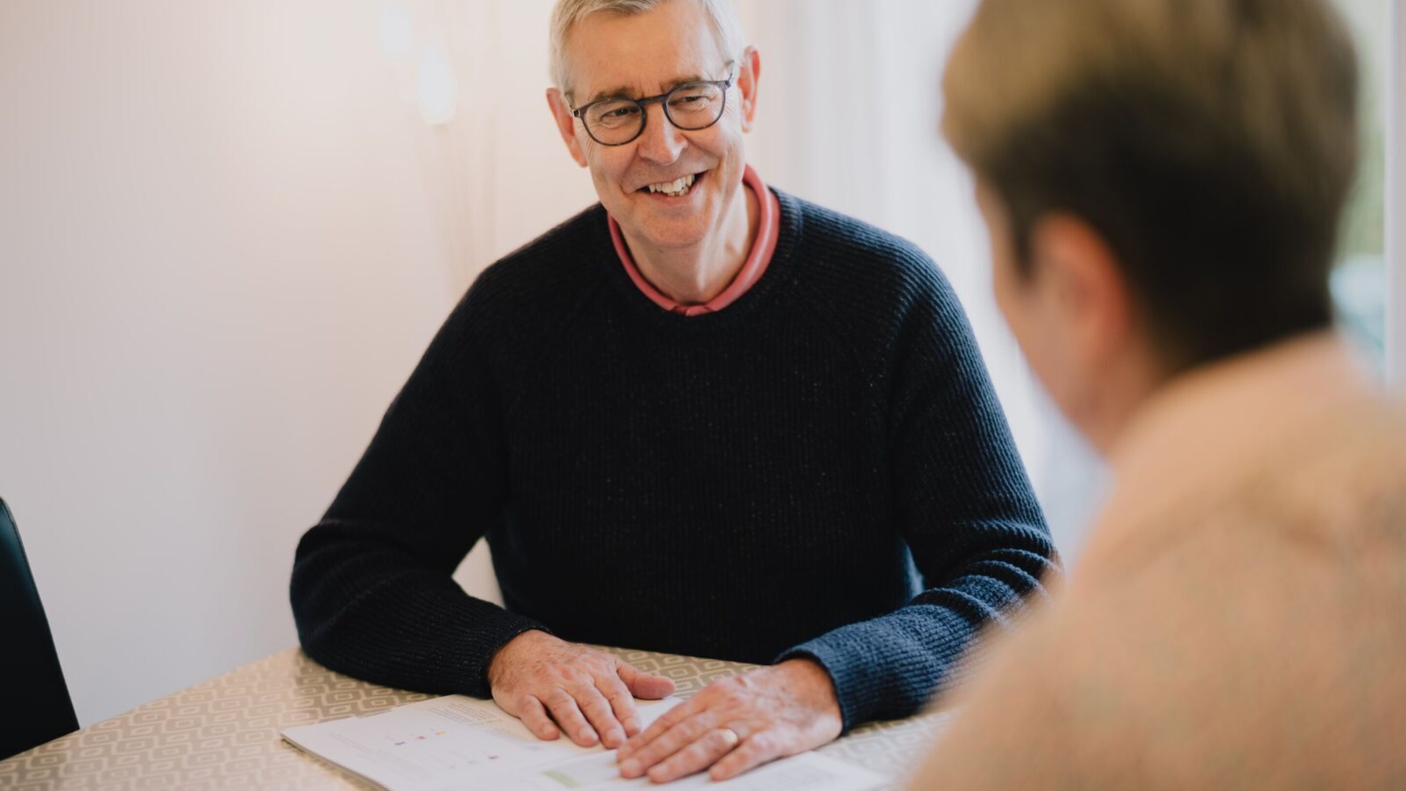 Retired CAP debt coach, Steve, sat at a debt client's table, holding paperwork and smiling