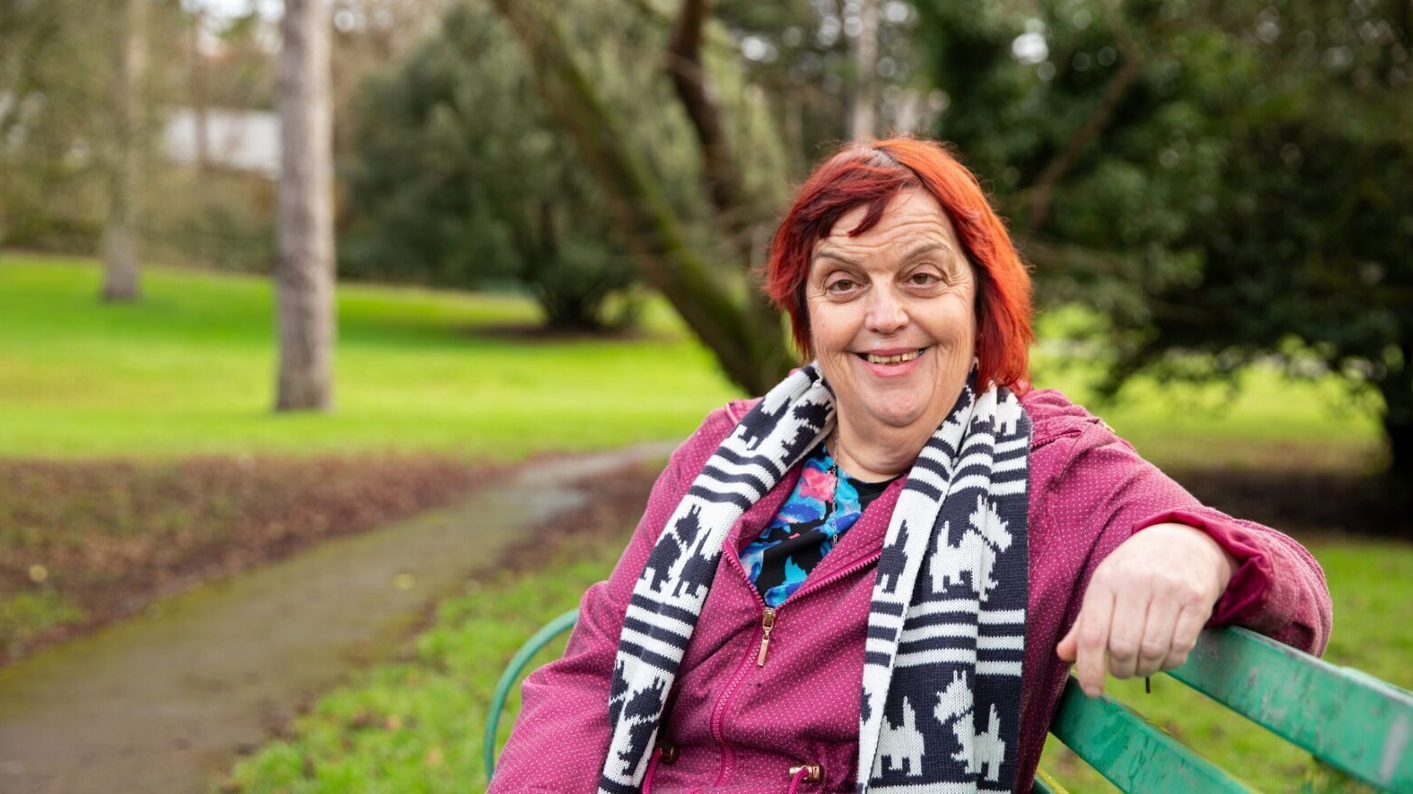 CAP client Julie, sat on a park bench and wearing a pink coat, smiling at the camera