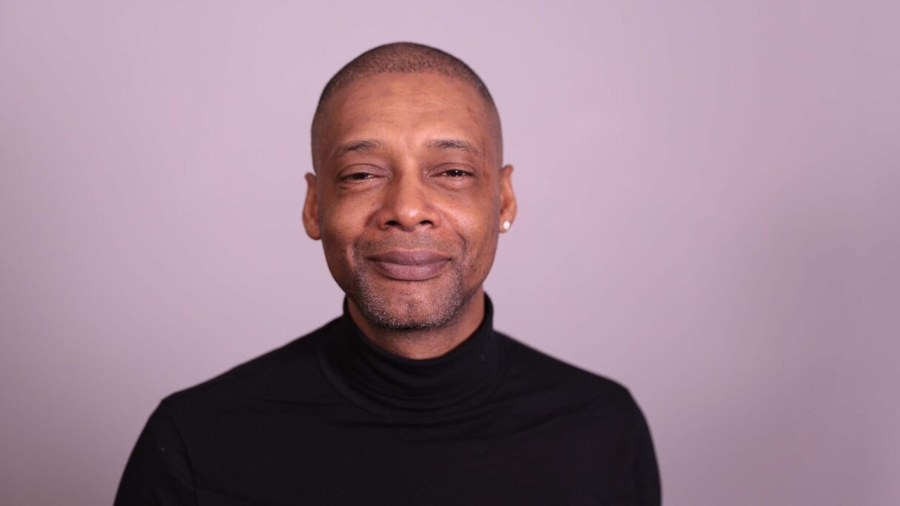 CAP client, Paul, stood against a purple wall, smiling at the camera and wearing a black turtleneck jumper