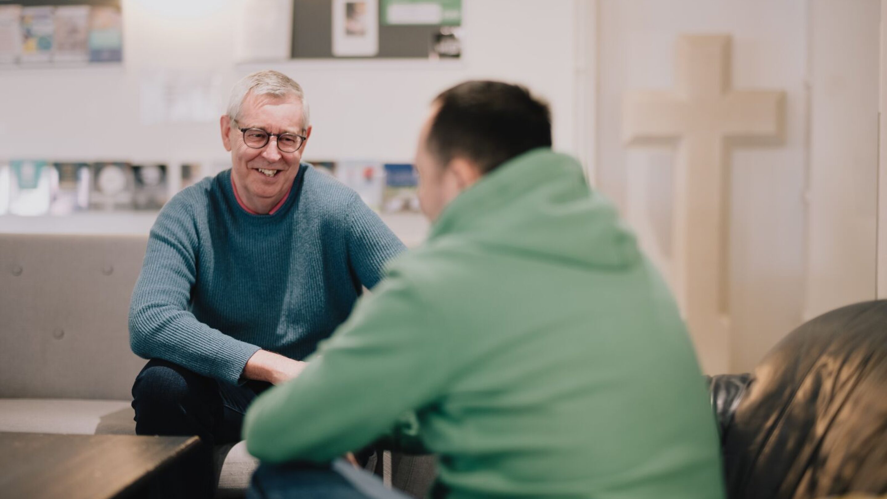 CAP debt coach, Steve, sat in a church with a CAP client, smiling at them. There is a large white cross in the background.