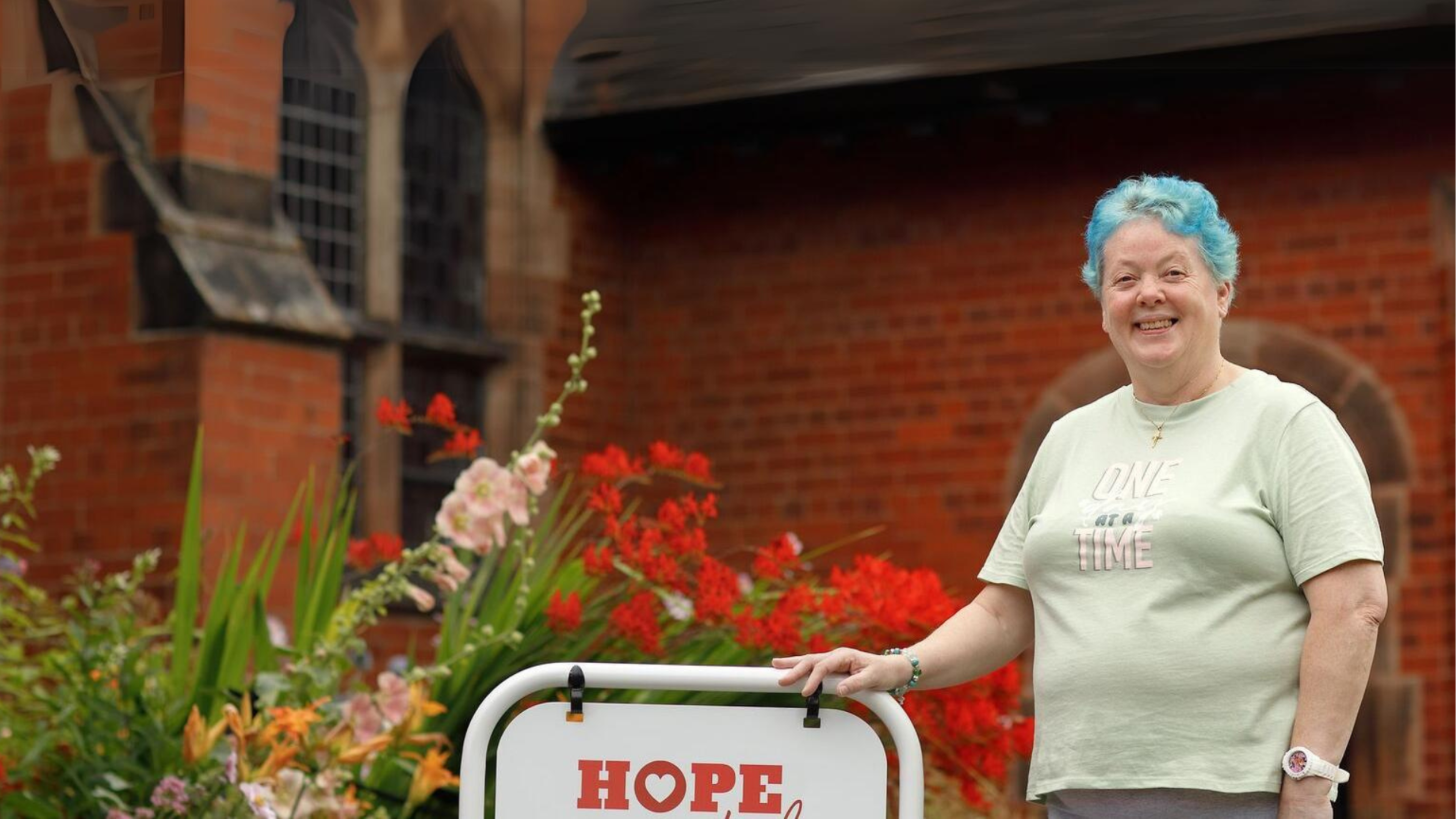 Bea, a lady with bright blue short hair, smiling at the camera. She's standing outside her church, holding on to a sign that reads 'hope' - the sign that led her to go into the church and start her journey to becoming debt free.