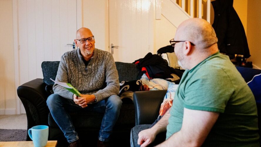 CAP client, Stevie, sat with his CAP debt coach in his living room. They are both looking at each other and smiling