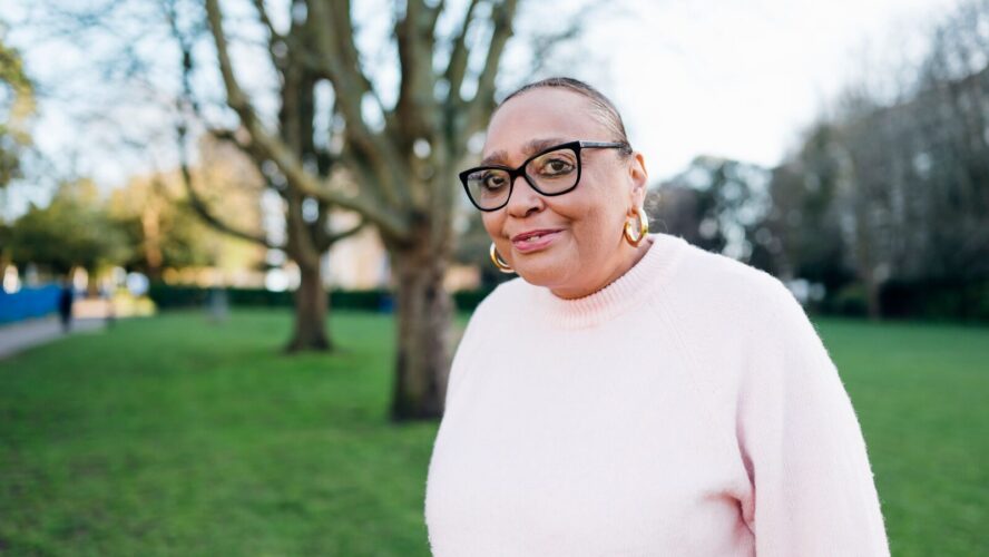 CAP client, Sharon, outside in a park, smiling at the camera and wearing a pink jumper.