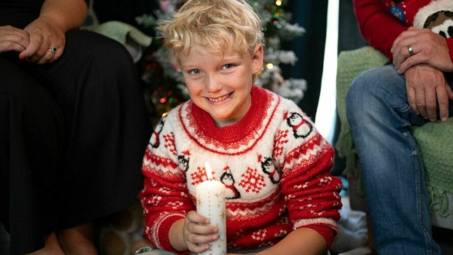 Image of a boy smiling and wearing a Christmas jumper, sat in front of the Christmas tree and holding a candle