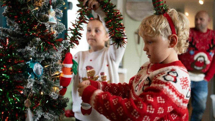 Two children wearing Christmas jumpers and antlers, decorating a Christmas tree while their dad watches on from across the room.