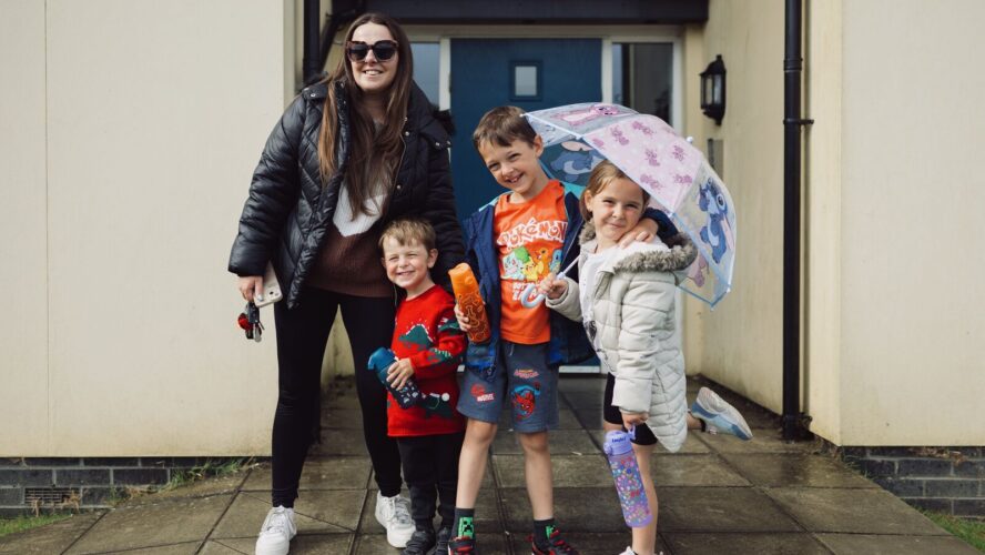 Family of four showing a mum and three children stood outside their home, smiling at the camera
