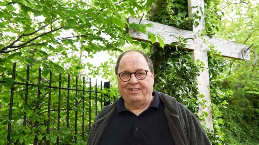 CAP client, Simon, stood outside in a garden, smiling, with a large wooden cross propped up against a tree behind him