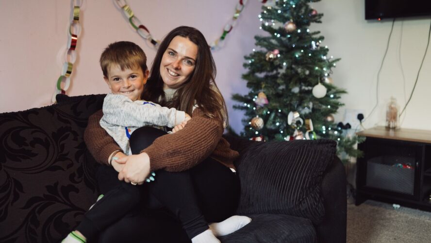 CAP client, Jade, sat on her sofa cuddling her son and smiling, with Christmas decorations and a Christmas tree behind them .