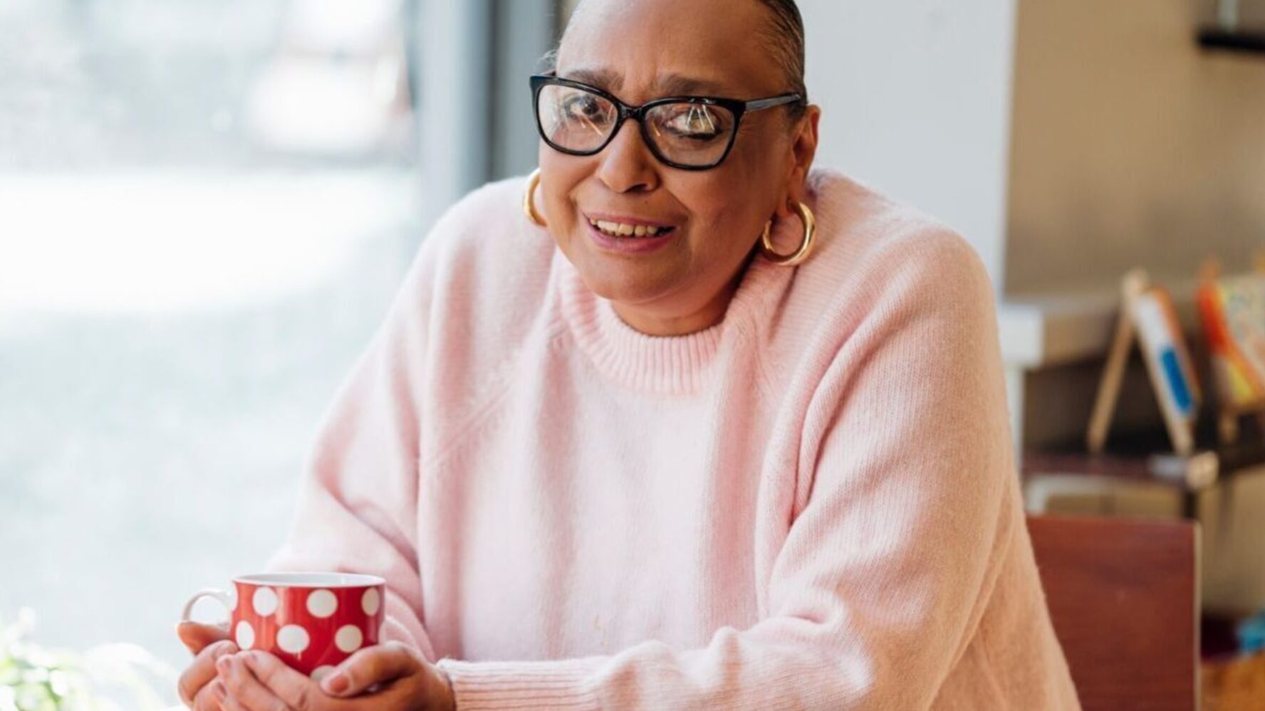 CAP client, Sharon, sat at a table holding a red mug, wearing a pink jumper and smiling at the camera