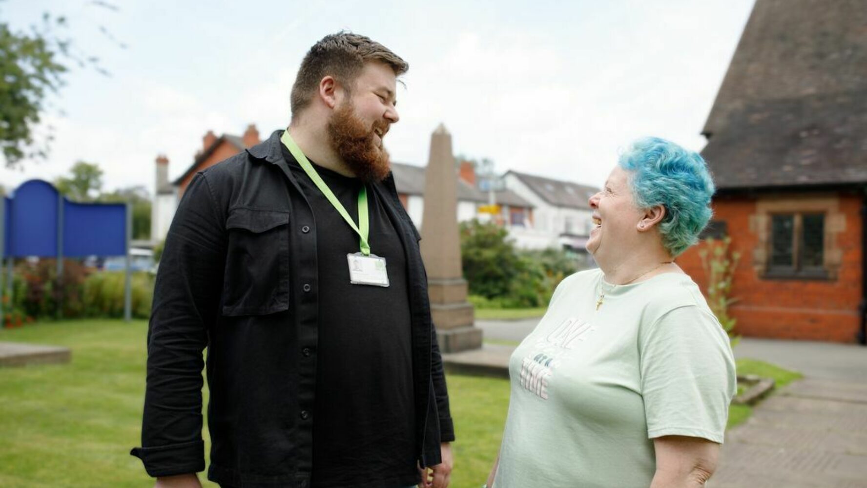 Outside a church. On the left is Barney, a CAP Debt Coach, with short brown hair, a beard and checked shirt. On the right is Bea, now debt free, who has bright blue short hair and a t-shirt. They're looking at each other and laughing happily.